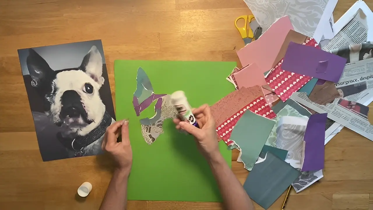 Hands are crafting a collage of a dog using glue and cut-out colored paper pieces on a green sheet. A photo of a dog is visible on the left. Scissors, paper scraps, and glue are scattered around.
