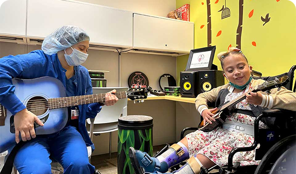 In a brightly decorated room, a person in scrubs plays guitar as part of sound healing therapy, while a young girl in a wheelchair, wearing braces, joyfully strums her ukulele.