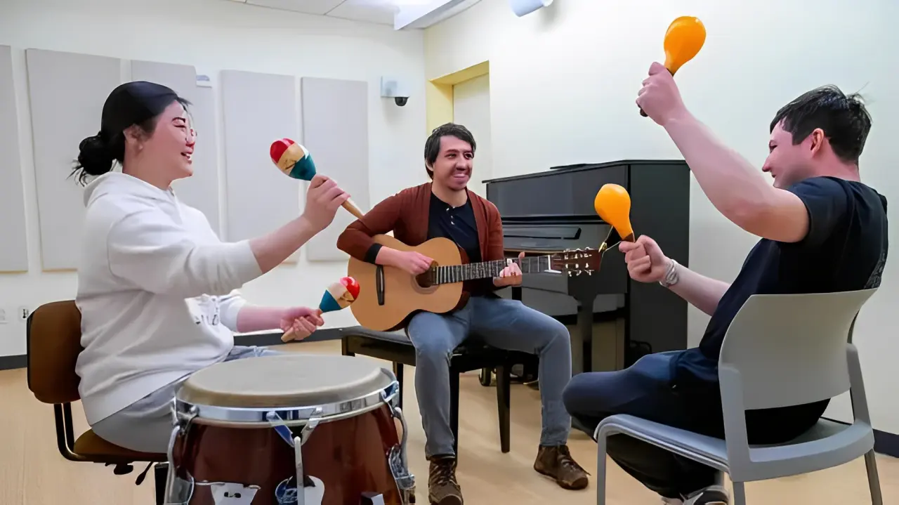 Three people in a music room are playing instruments, creating a vibrant rhythm. One with maracas, another with a drum, and the third with a guitar—each engaged as if conducting their own sound healing session through harmonious performance.