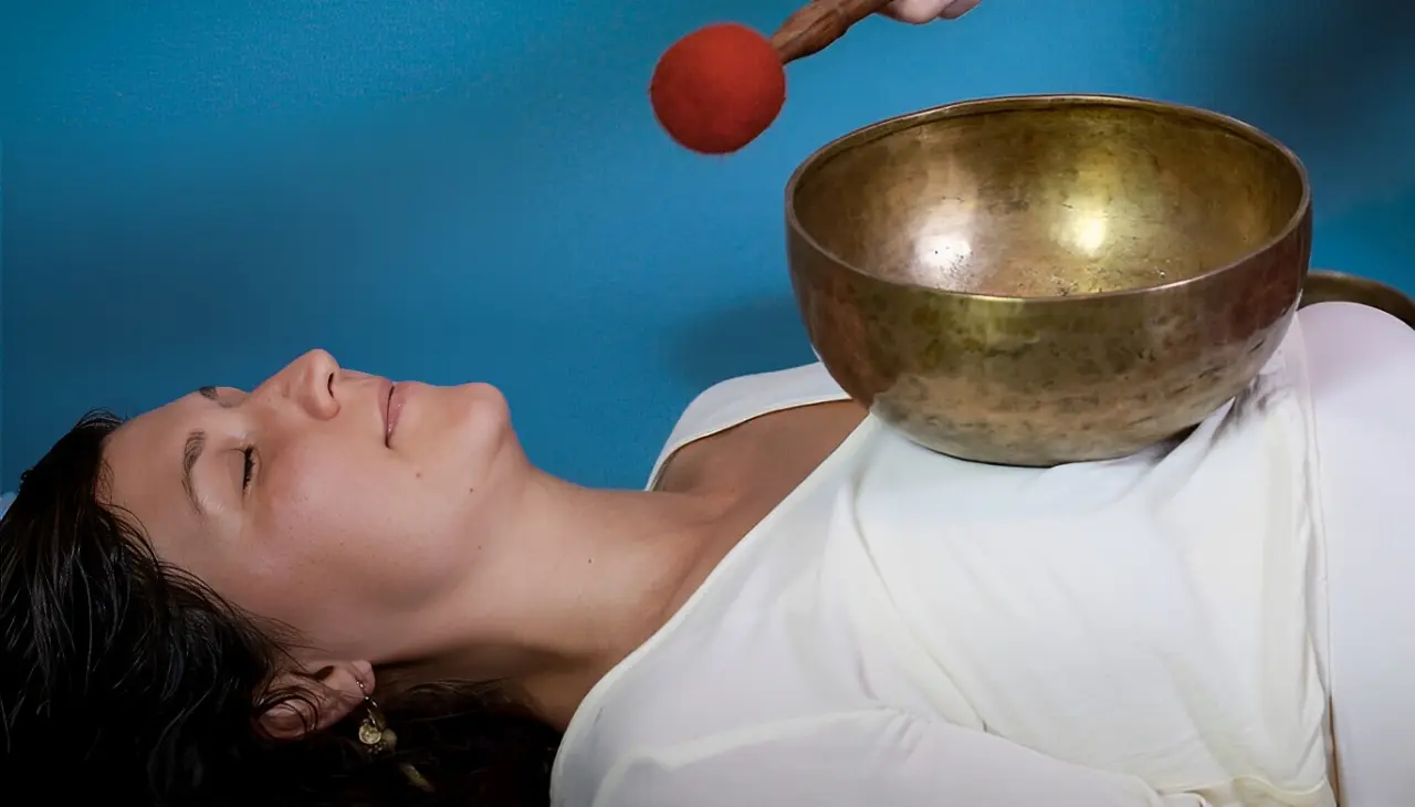 A person lies down with a singing bowl on their chest while another hand holds a mallet above it.