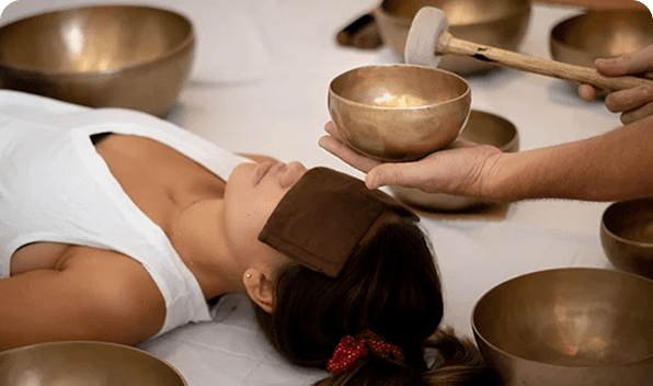 A person lies down with eyes gently covered, enveloped by the resonant presence of singing bowls. This serene scene is part of a sound healing therapy session, as another individual carefully prepares to strike a bowl with a mallet, ready to evoke soothing vibrations.