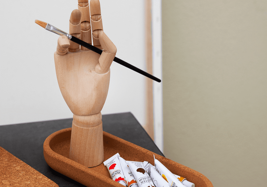 A wooden hand model holding a paintbrush above a tray with paint tubes.